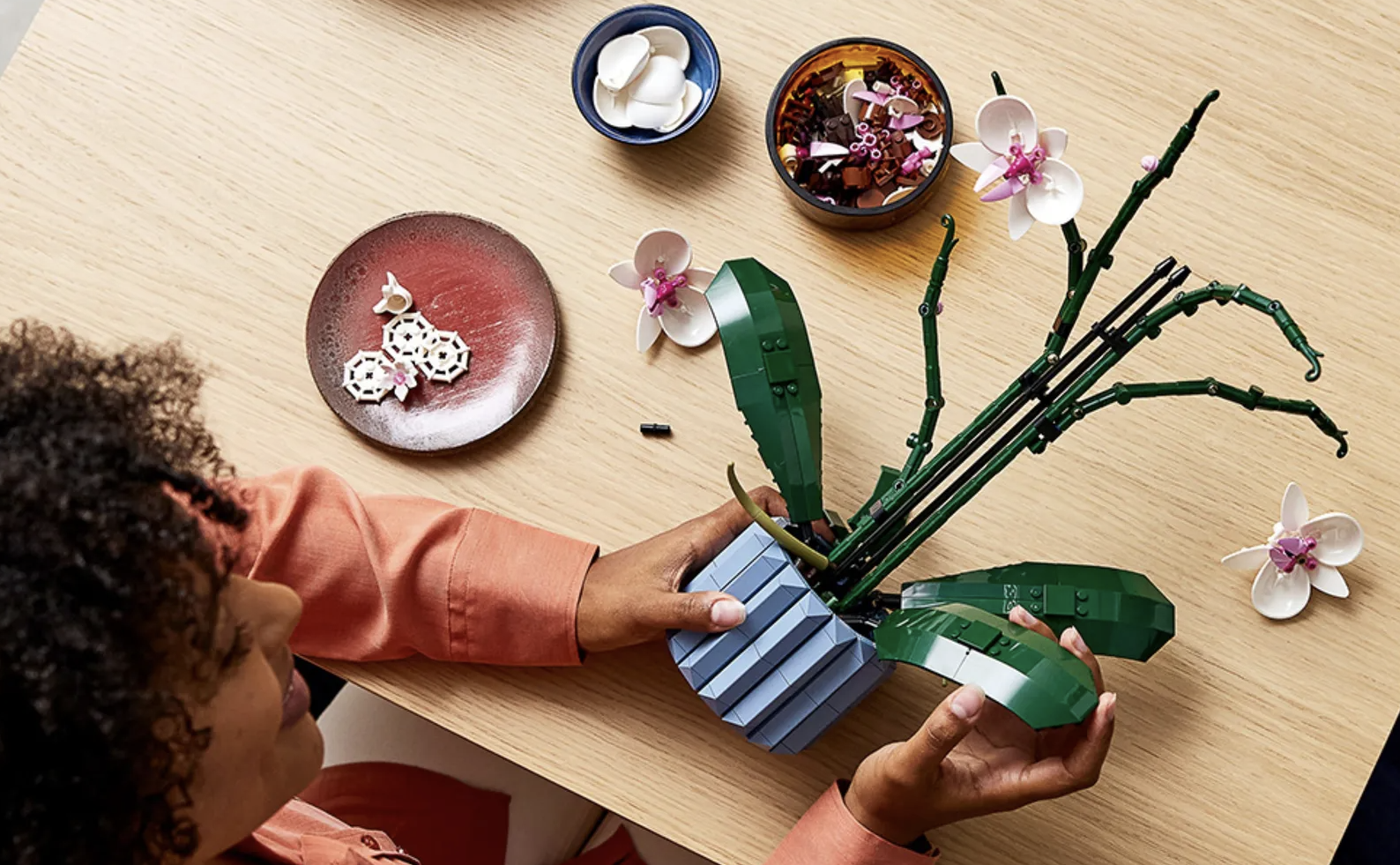 a woman in an orange shirt assembles the lego orchid kit on a wooden table