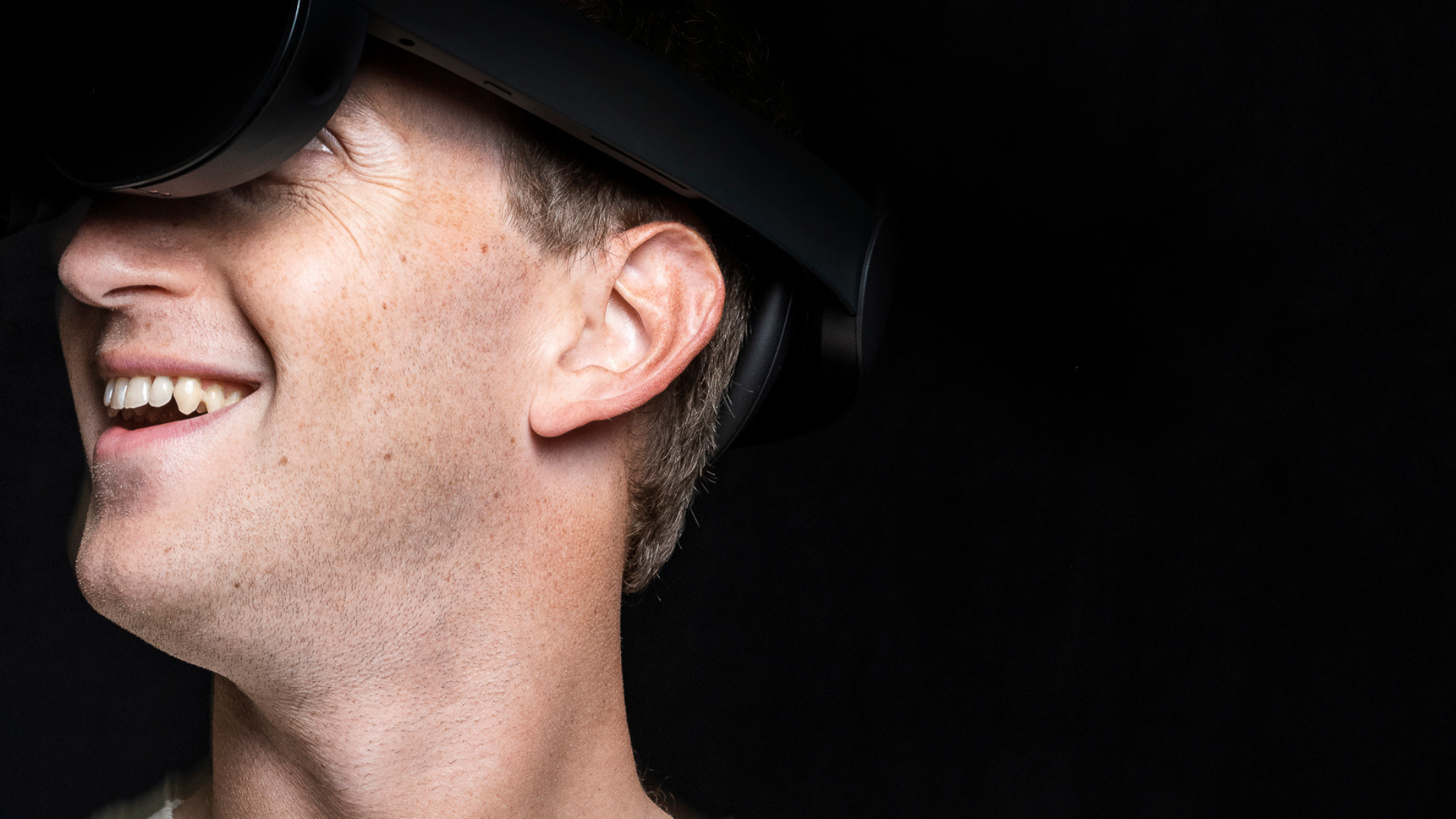 Mark Zuckerberg smiles into a holocake headset (basically, a flatter version of the headsets currently available) against a black background.)