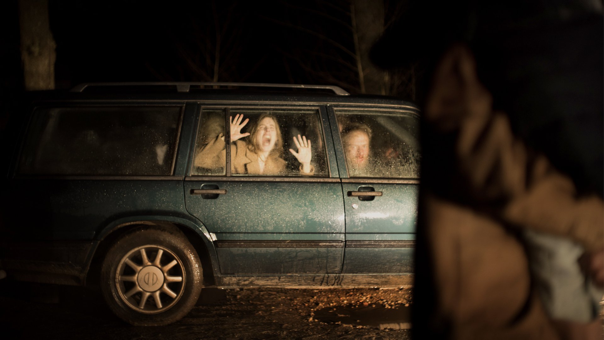 A woman screams in the backseat of a car, pressing her hands against the window and looking out at someone in the darkness.