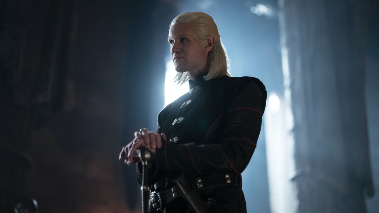 Matt Smith as Daemon Targaryen, a white man with white-blond hair resting his hands on the pommel of a sword.