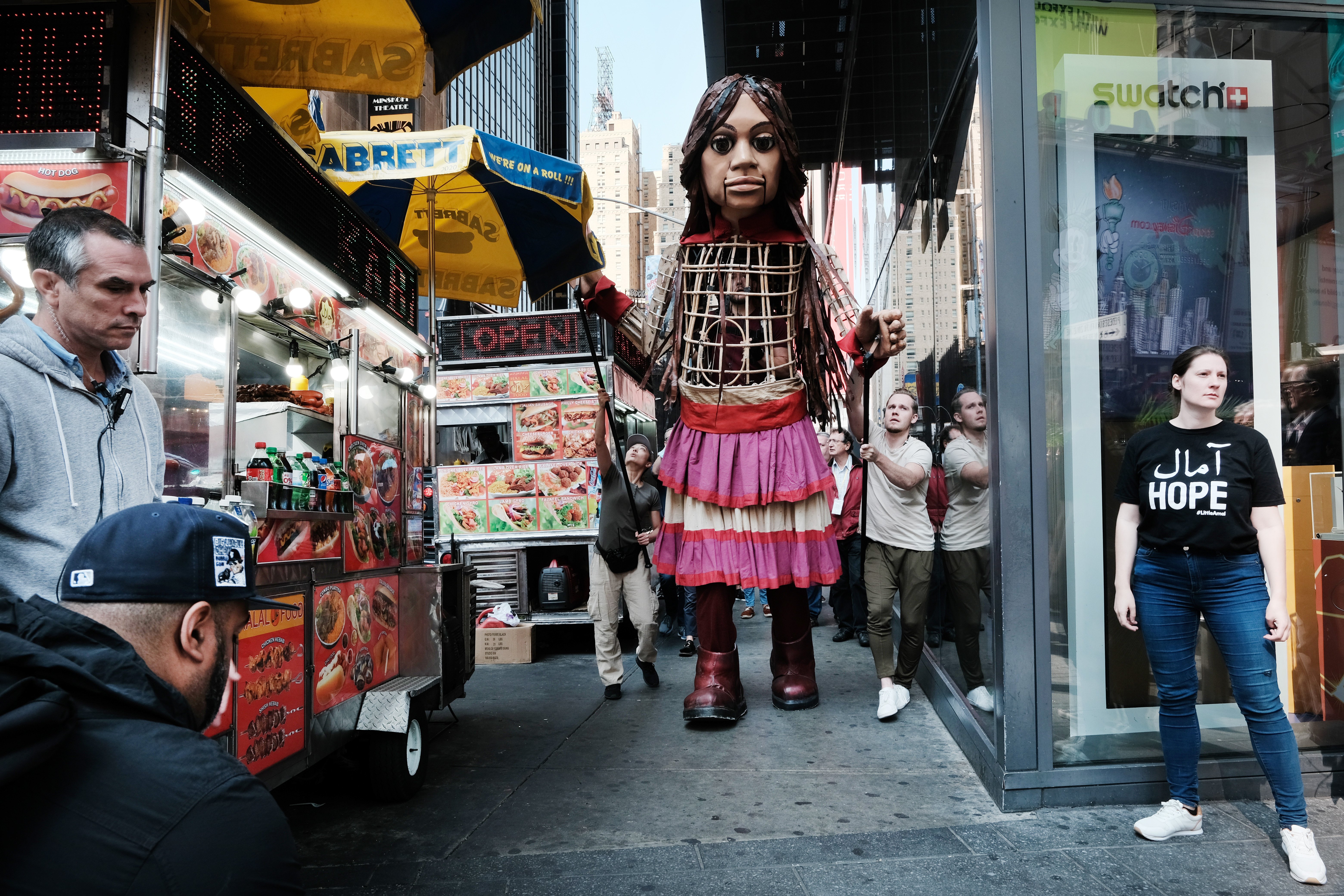 Amal walks through a busy New York City street, passing a hot dog stand and a subway entrance.