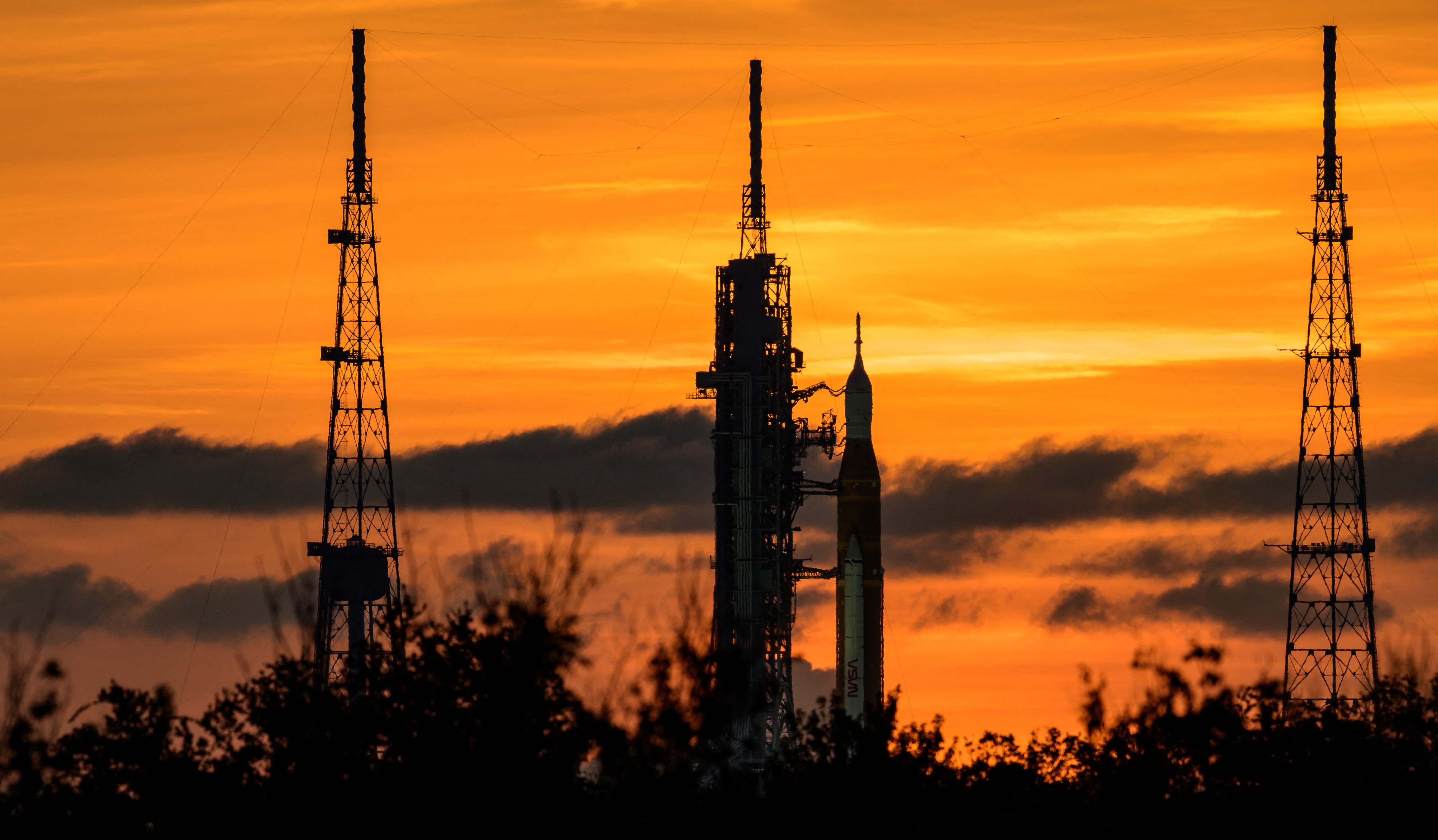 NASA's Space Launch System (SLS) rocket with the Orion spacecraft aboard — also known as Artemis I —sits on the launch pad on Aug. 31 ahead of its planned (and later cancelled) Sep. 3 launch. The sunset behind it is made of deep and bright oranges, and you can really only see the black outline of the rocket and system against it.