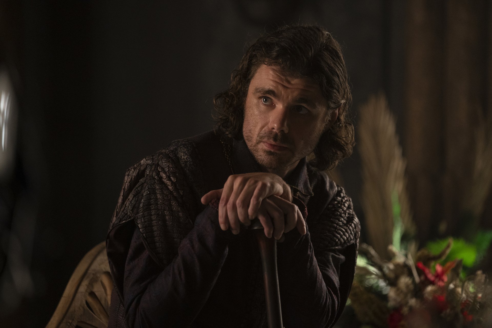 Matthew Needham, a white man with wavy, chin-length brown hair leaning on a wooden cane, as Ser Larys Strong in House of the Dragon.