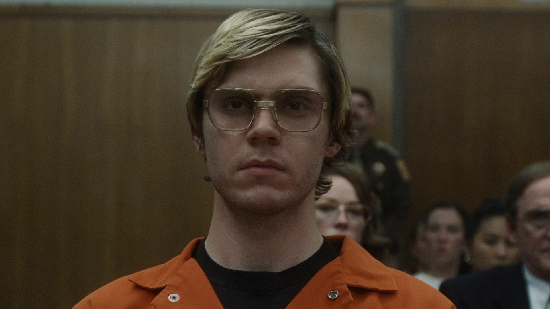 A man with glasses in an orange jumpsuit stands in a courtroom.