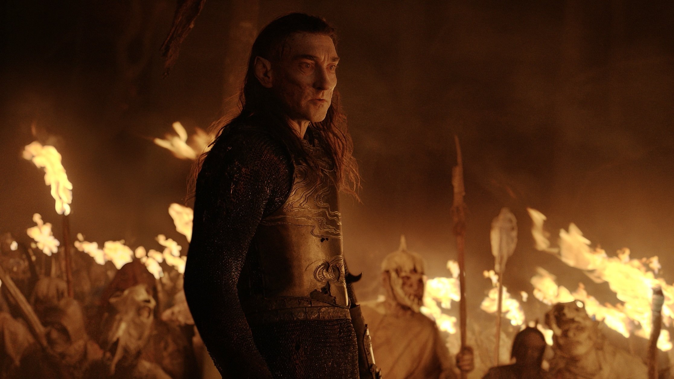 An elf with extremely pale and pockmarked skin stands, armored, in front of a horde of orcs carrying torches.