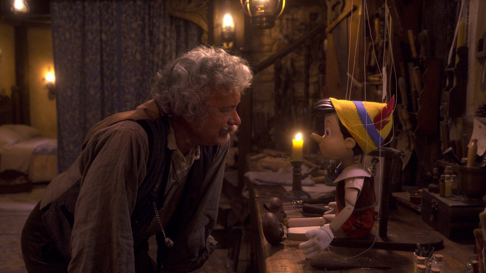 A man with a grey mustache stares at a wooden marionette puppet in his workshop.