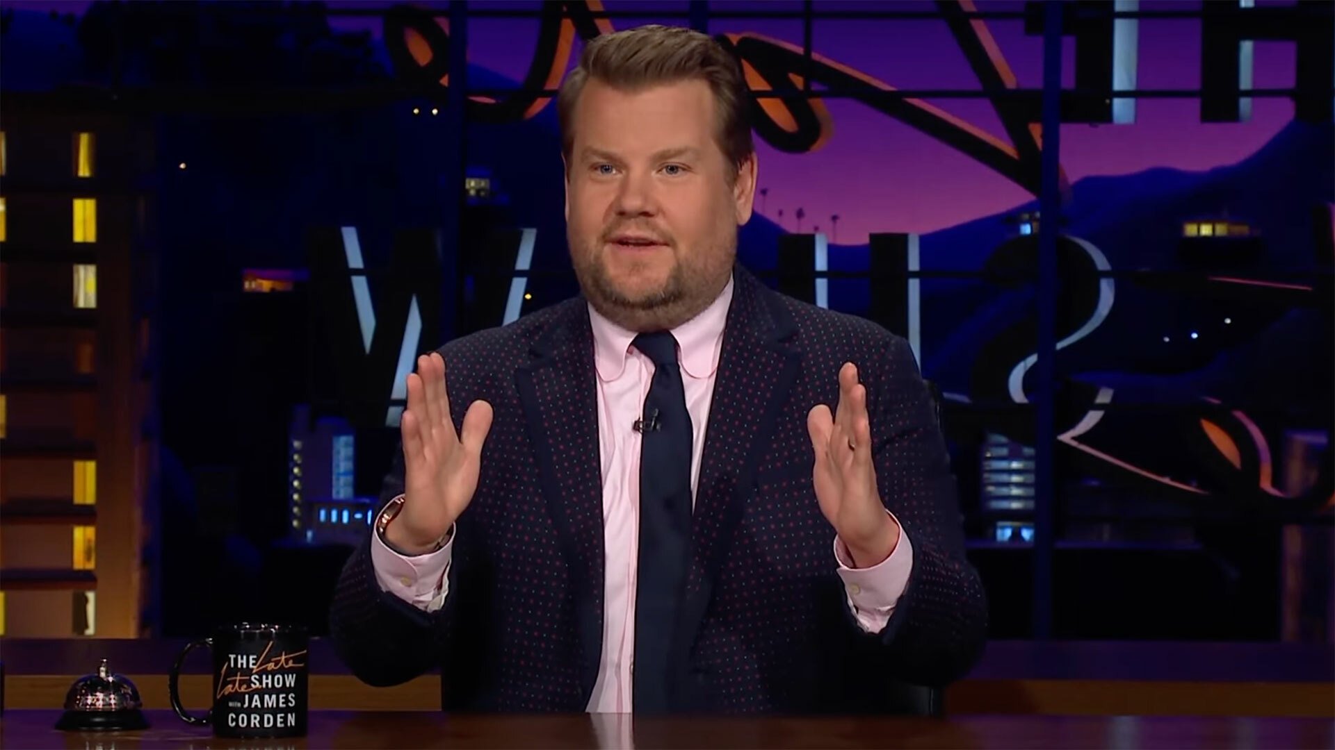 A man sitting behind a desk in a suit gestures with his hands.
