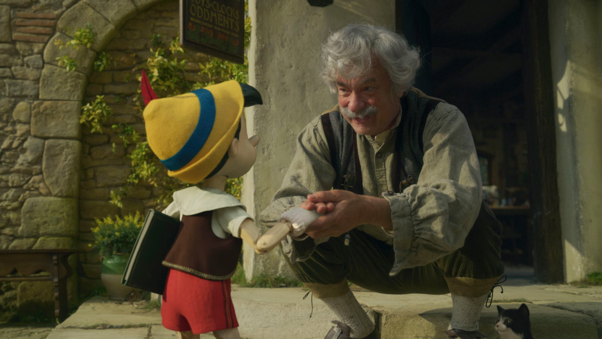 A man with grey hair and a mustache holds the hand of a wooden puppet boy, smiling.