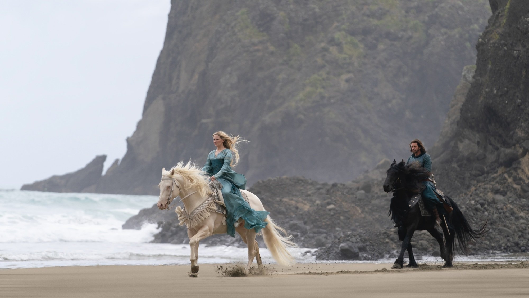 An elven women in a blue dress rides down a beach on a white horse. A human man in blue armor on a black horse follows her.