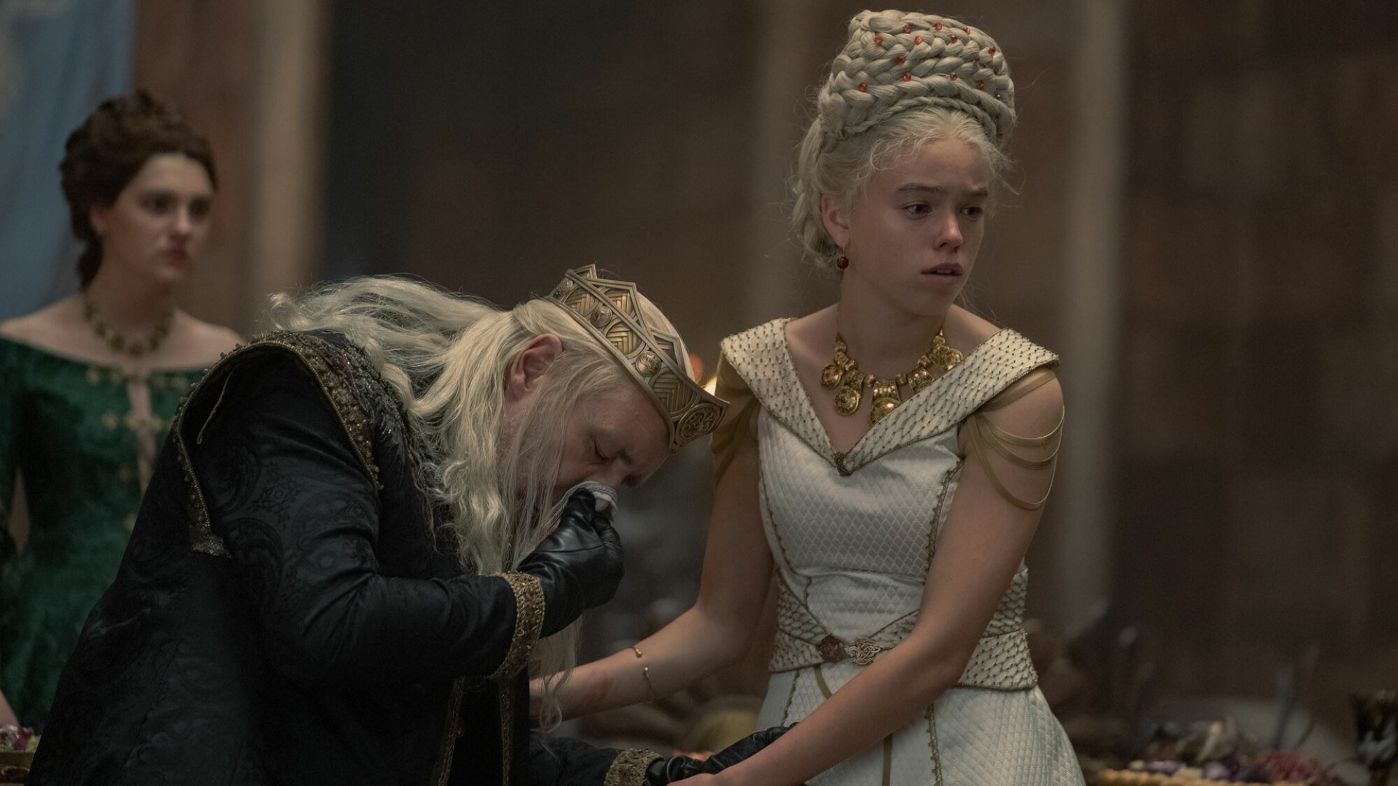 A king coughs into a handkerchief while a blonde woman holds onto him.