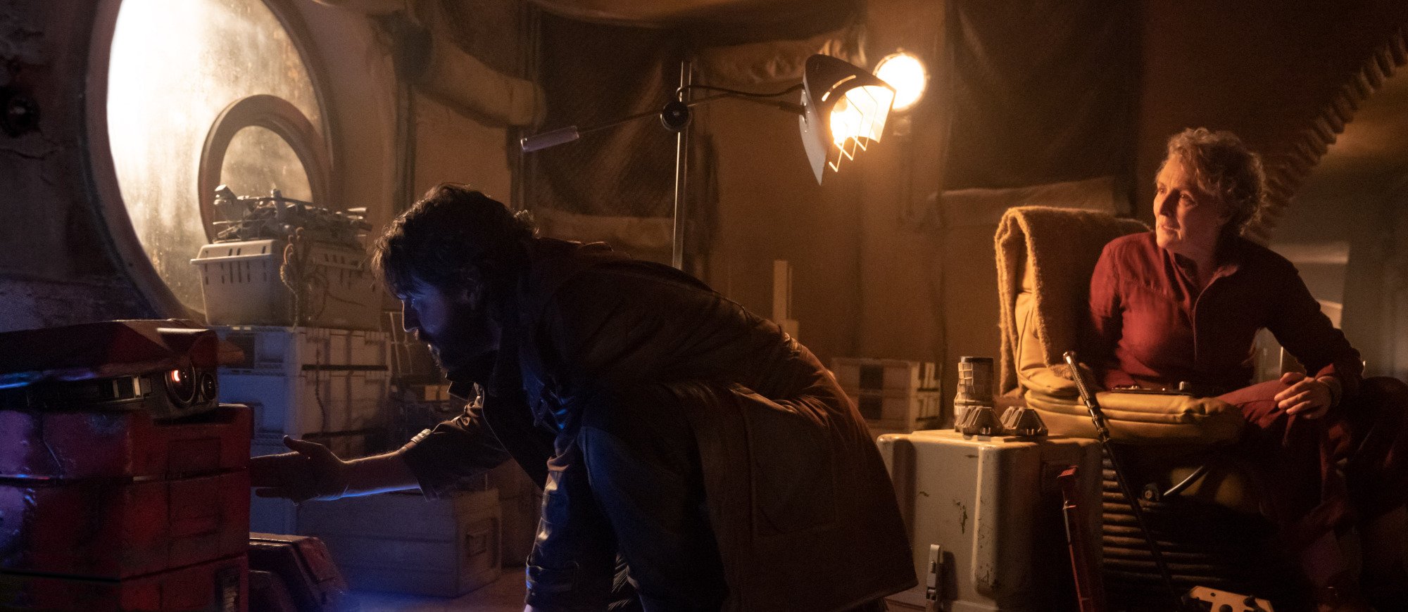 Cassian Andor crouches behind boxes while his stepmom looks at him.