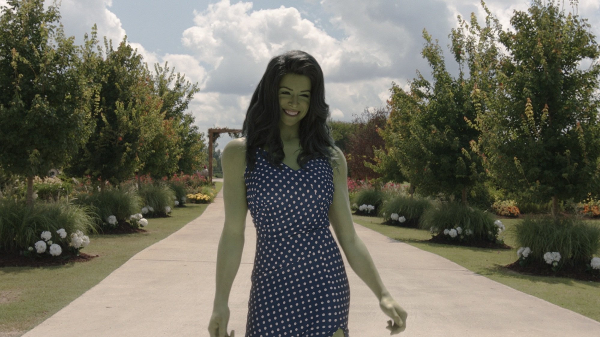 She-Hulk, a giant woman with green skin, walks down a dirt road lined with neatly trimmed trees.