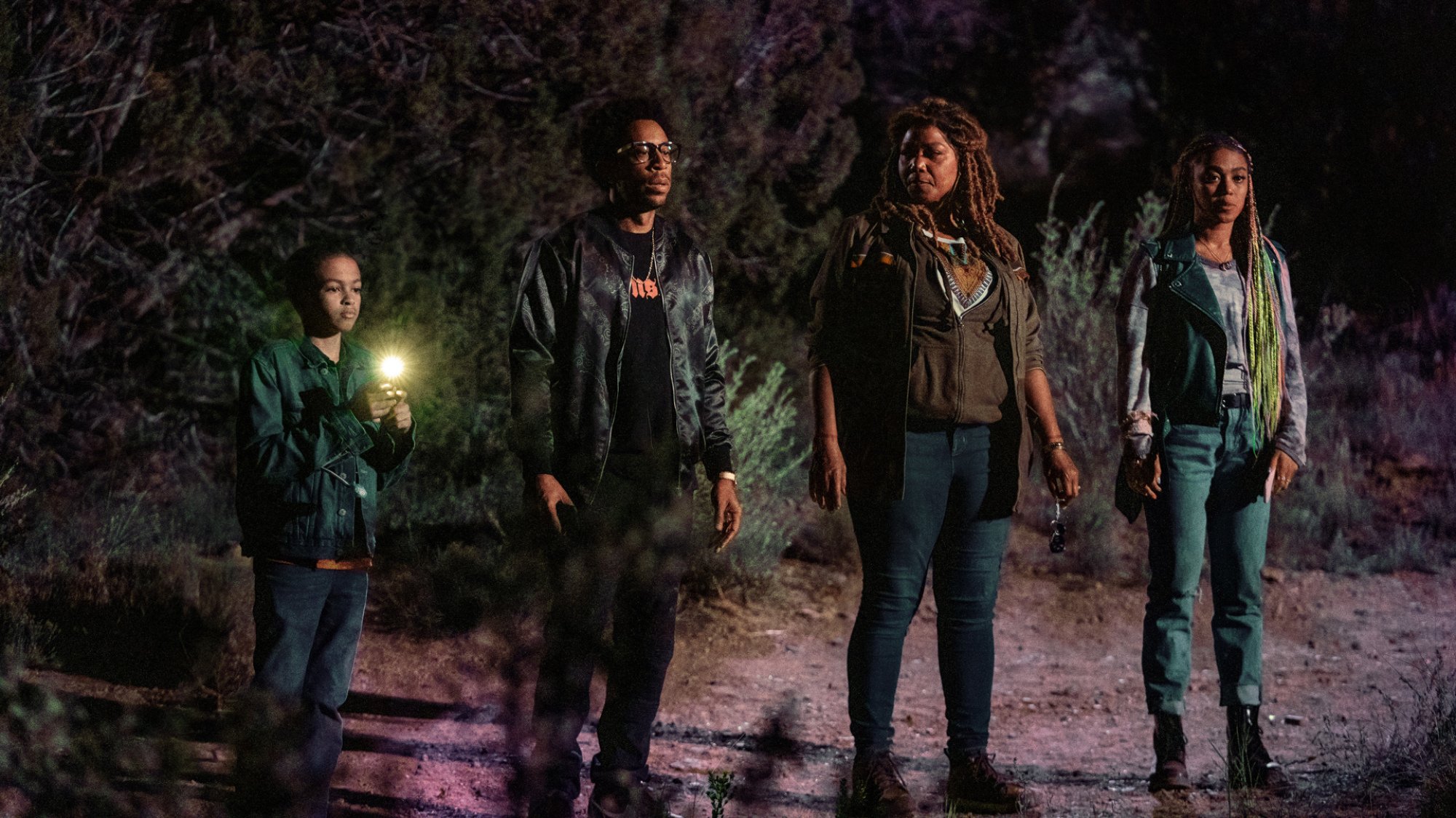 Four people stand in a line in the desert at night, one with a torch.