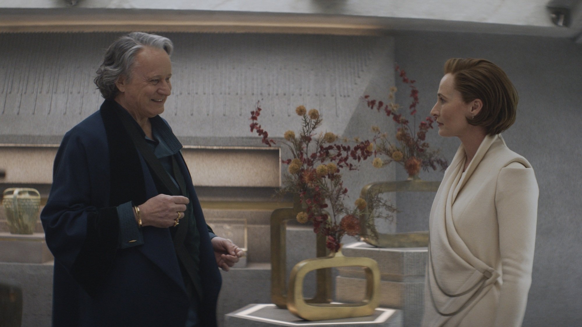 An older man and a woman talk in a stony apartment full of gold flower vases.