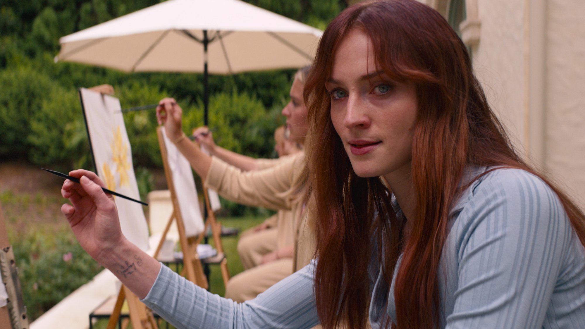 A girl sits with others painting outdoors, staring menacingly offscreen.