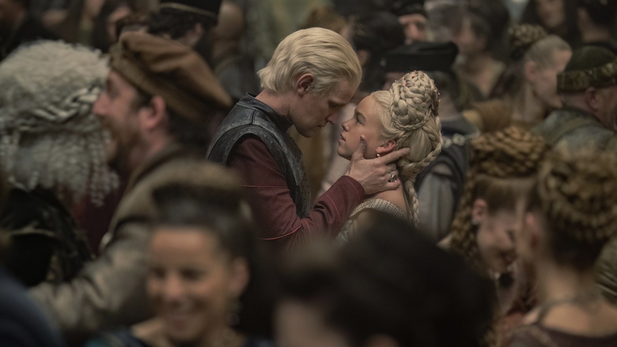 A man with silvery-blonde hair holds the face of a young woman with silvery blonde hair. They are in the middle of a crowded dancefloor.