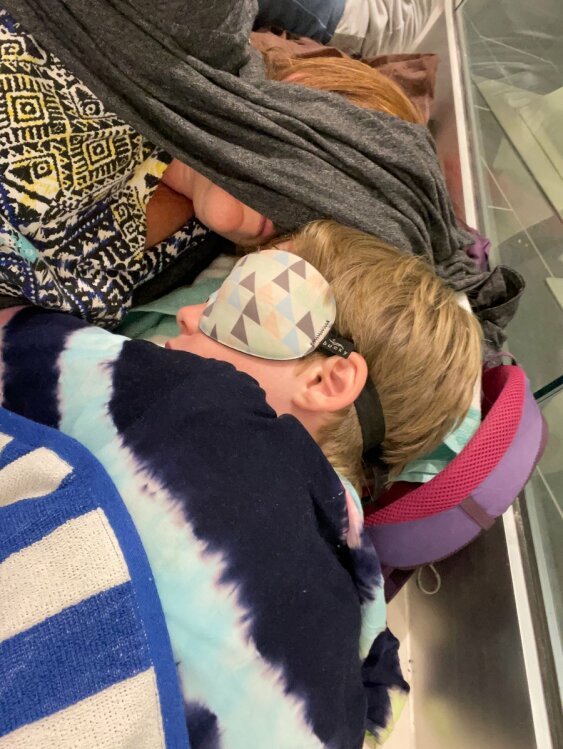 Henry sleeps on a travel pillow on the floor of the mall, his eyes covered by a sleep mask. His mom is lying next to him and using a sweater to cover her eyes.