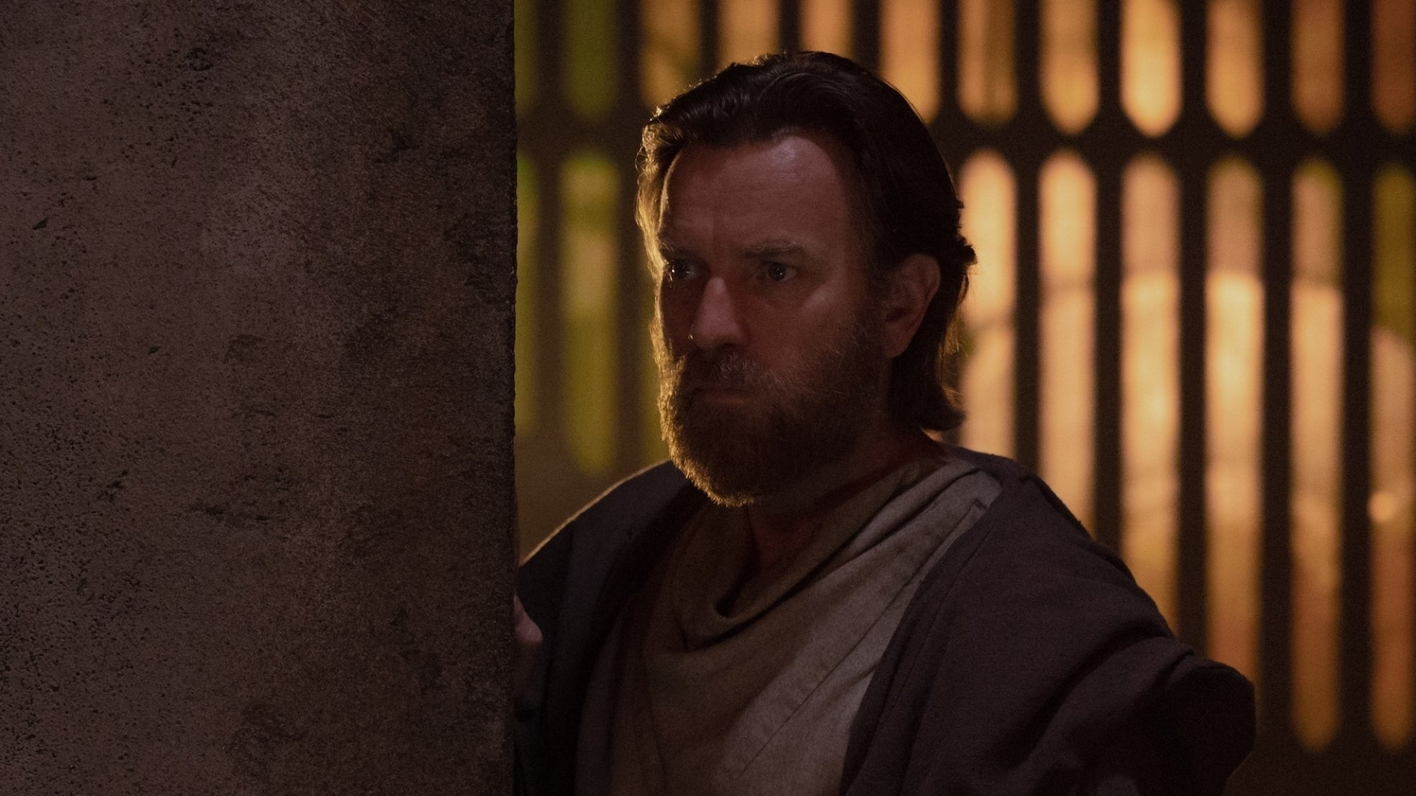A man in robes leans around a corner looking pensive.