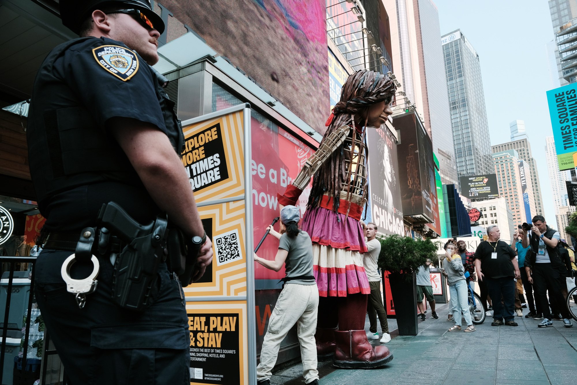 Amal leans against a building. An NYC police offer stands in the foreground with arms clasped in front of him.
