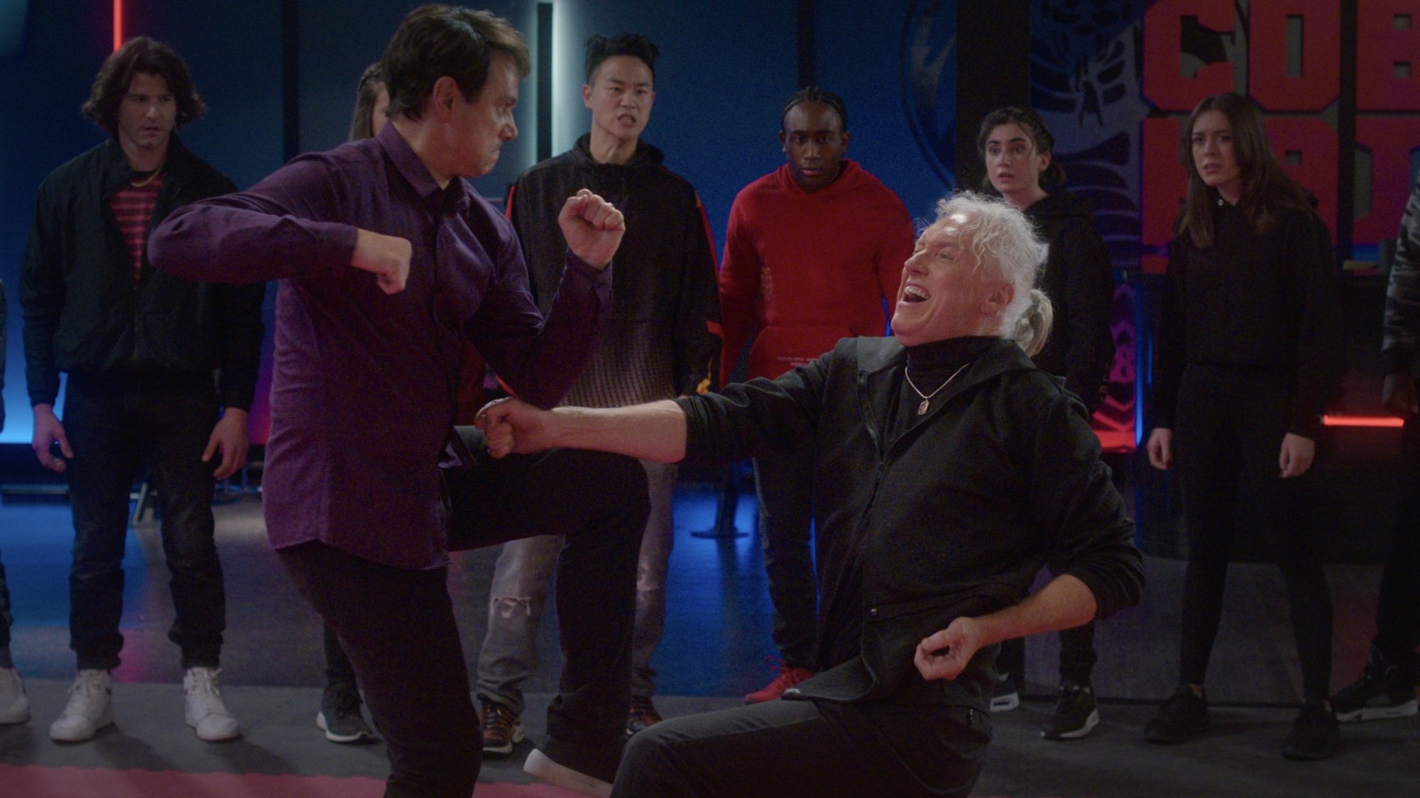 Two men engage in a karate fight as their students look on.