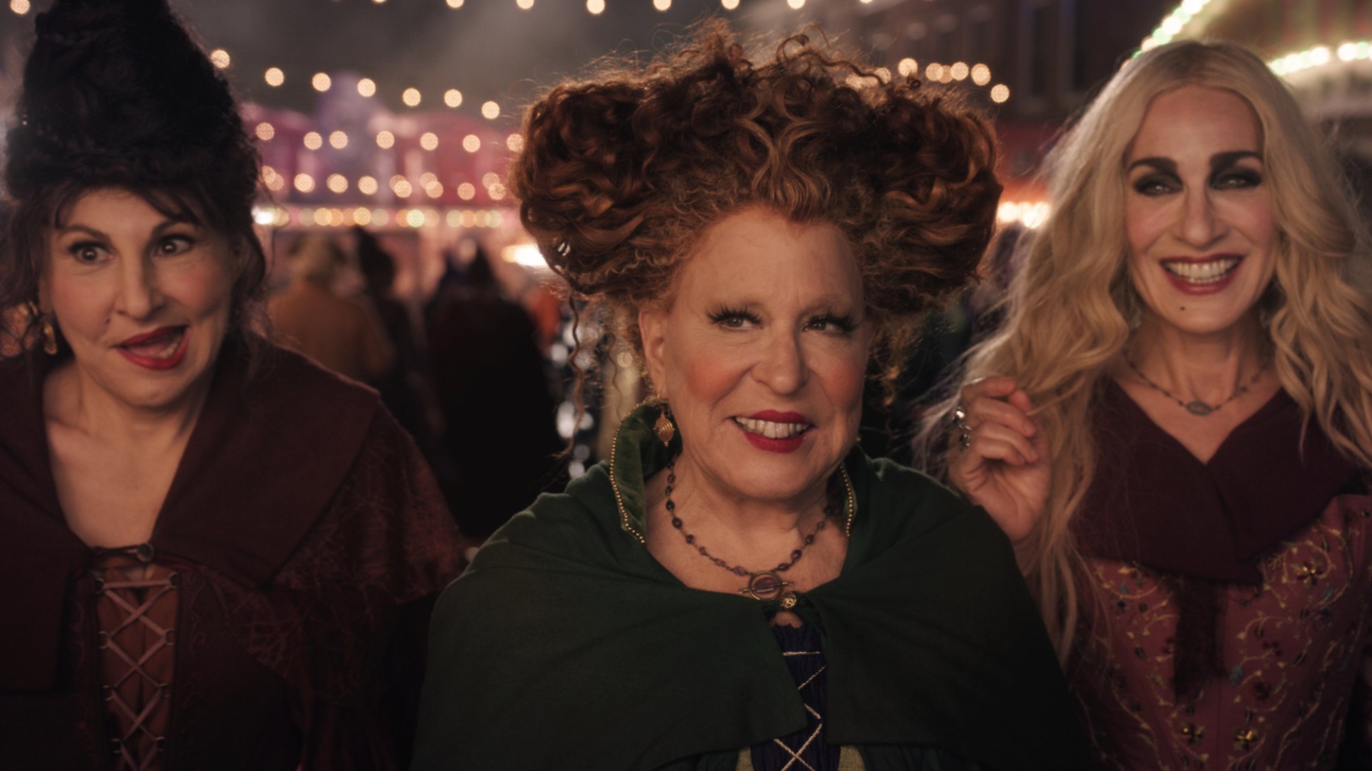 Three actors dressed as witches walk through a carnival, smiling.