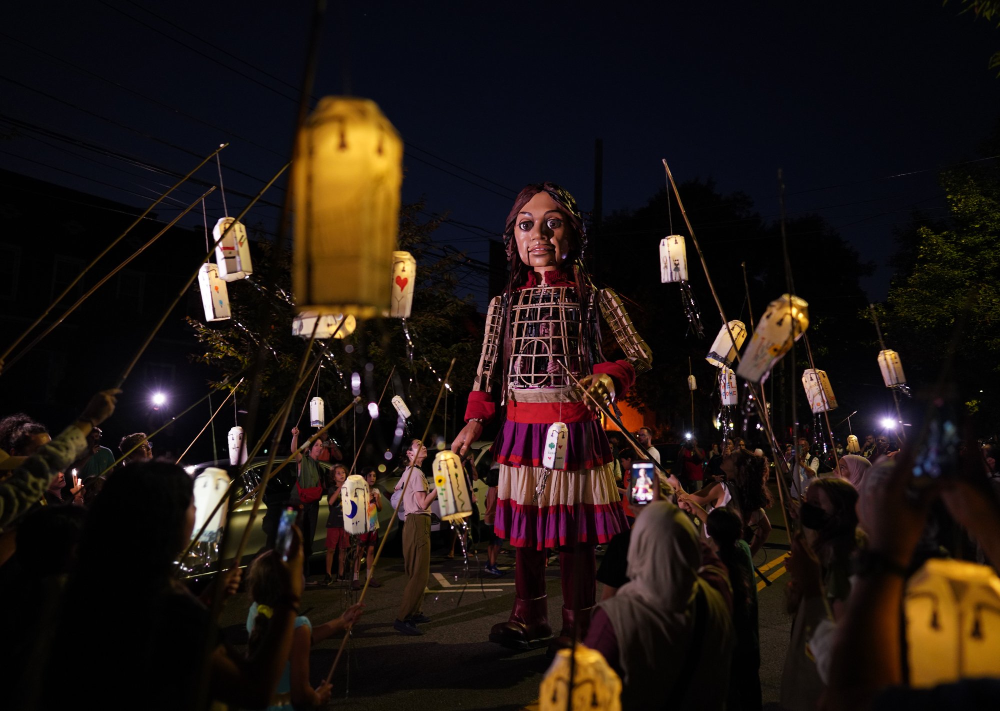Amal walks through a crowd during the night time, as individuals hold up many paper lanterns that glow around her.
