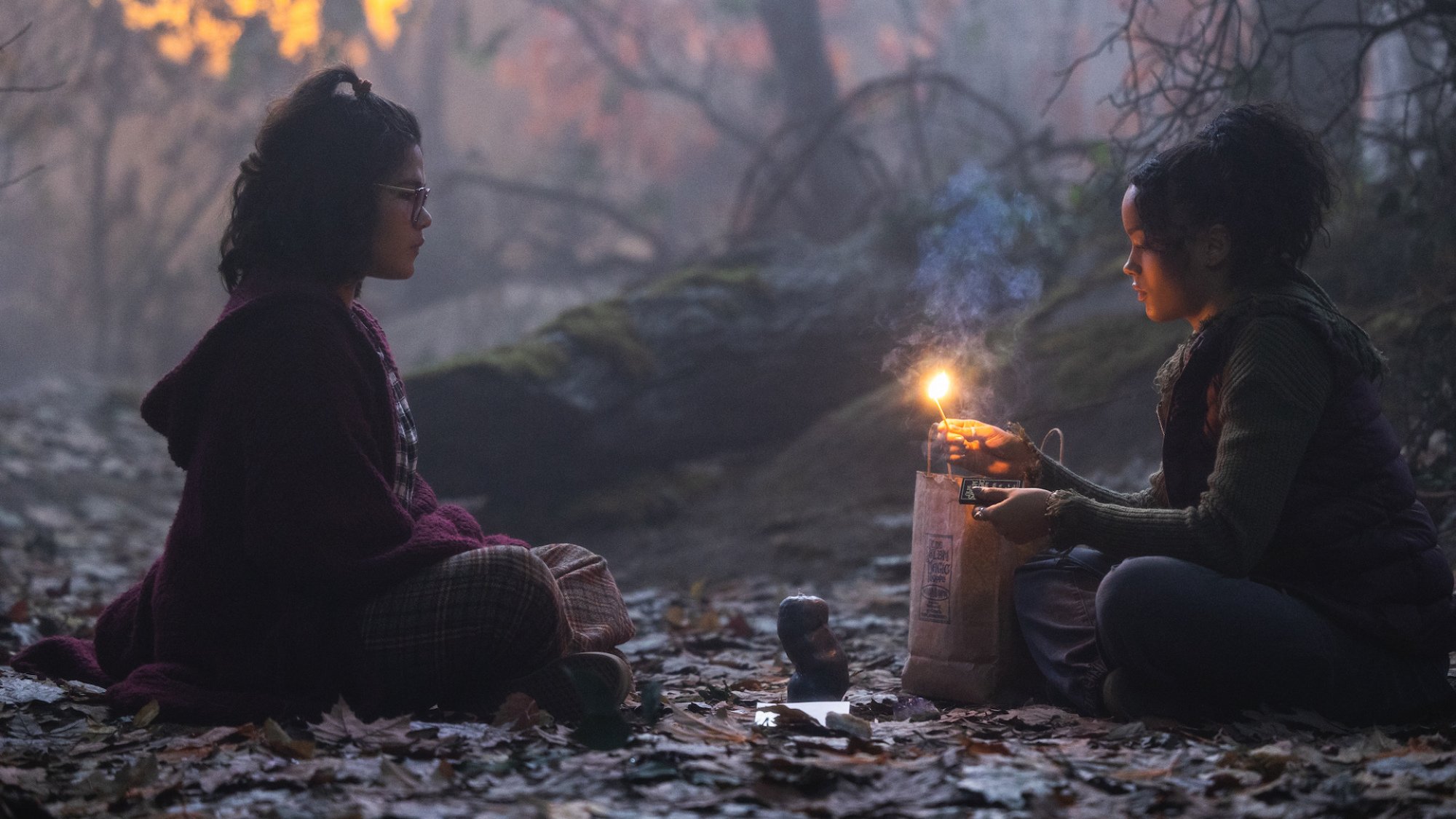 Two girls sit in a wood with a candle, facing each other.