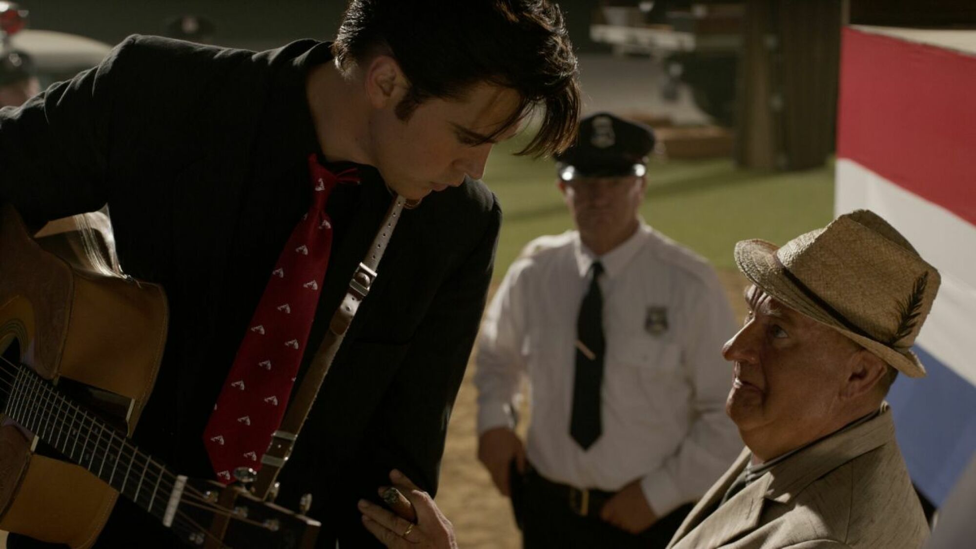 A man in a black suit with a guitar stands talking to a man in a white suit wearing a fedora.
