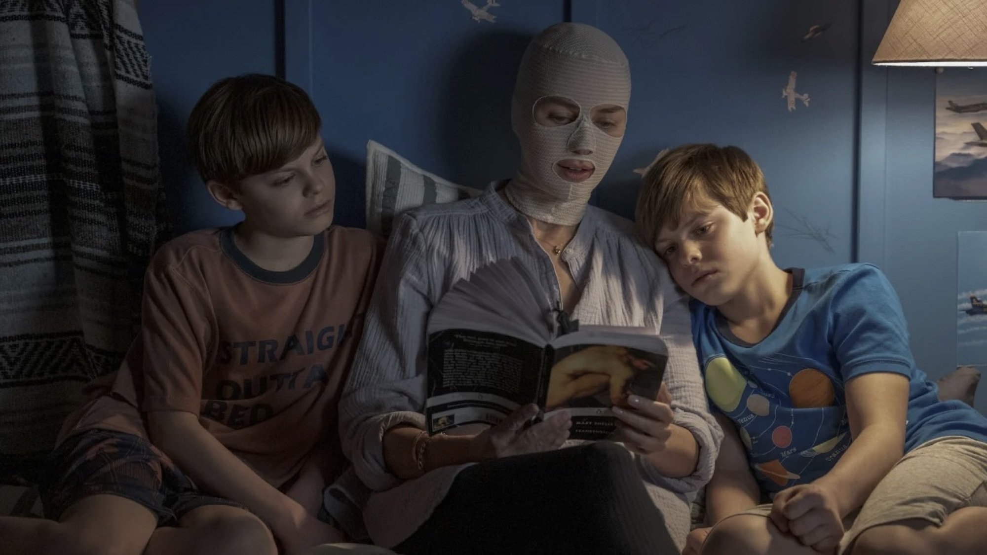 A woman with gauze wrapped around her head reads a story to her twin boys, who are in bed on either side of her.