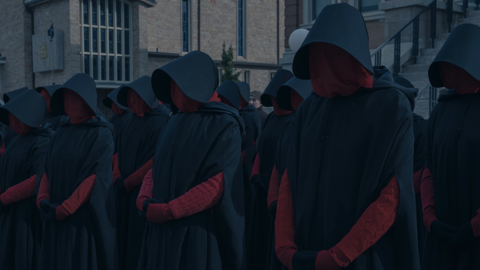 A group of black-cloaked women in red masks stand in an organized line.