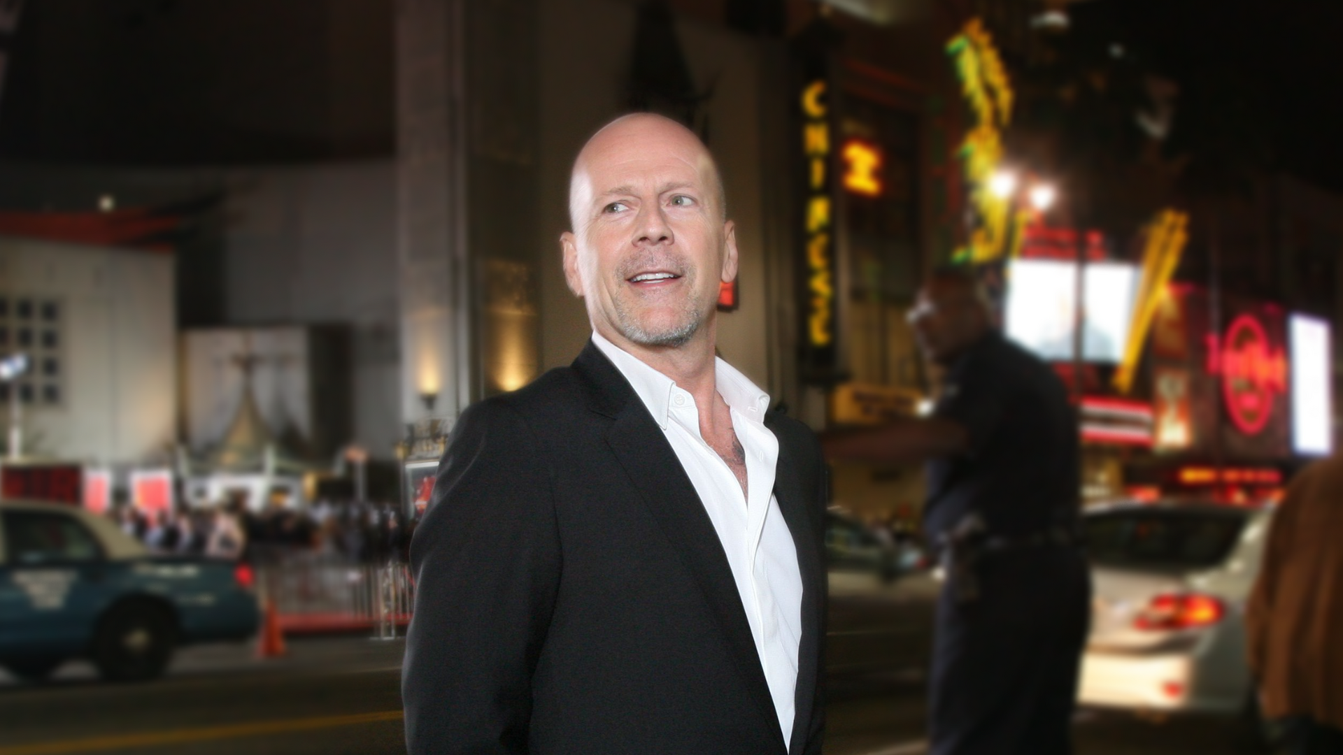 A photo of Bruce Willis from 2010 smiling and in a tuxedo on Hollywood Boulevard, across the street from Grauman's Chinese Theatre.