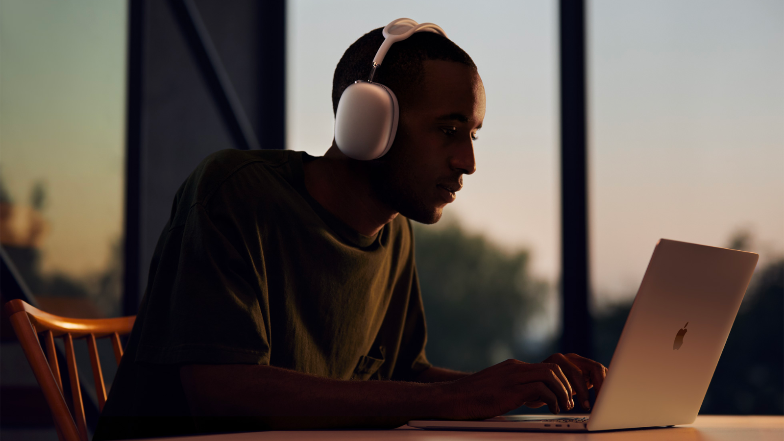 a man wearing silver airpods max working on a macbook in a room at sunset