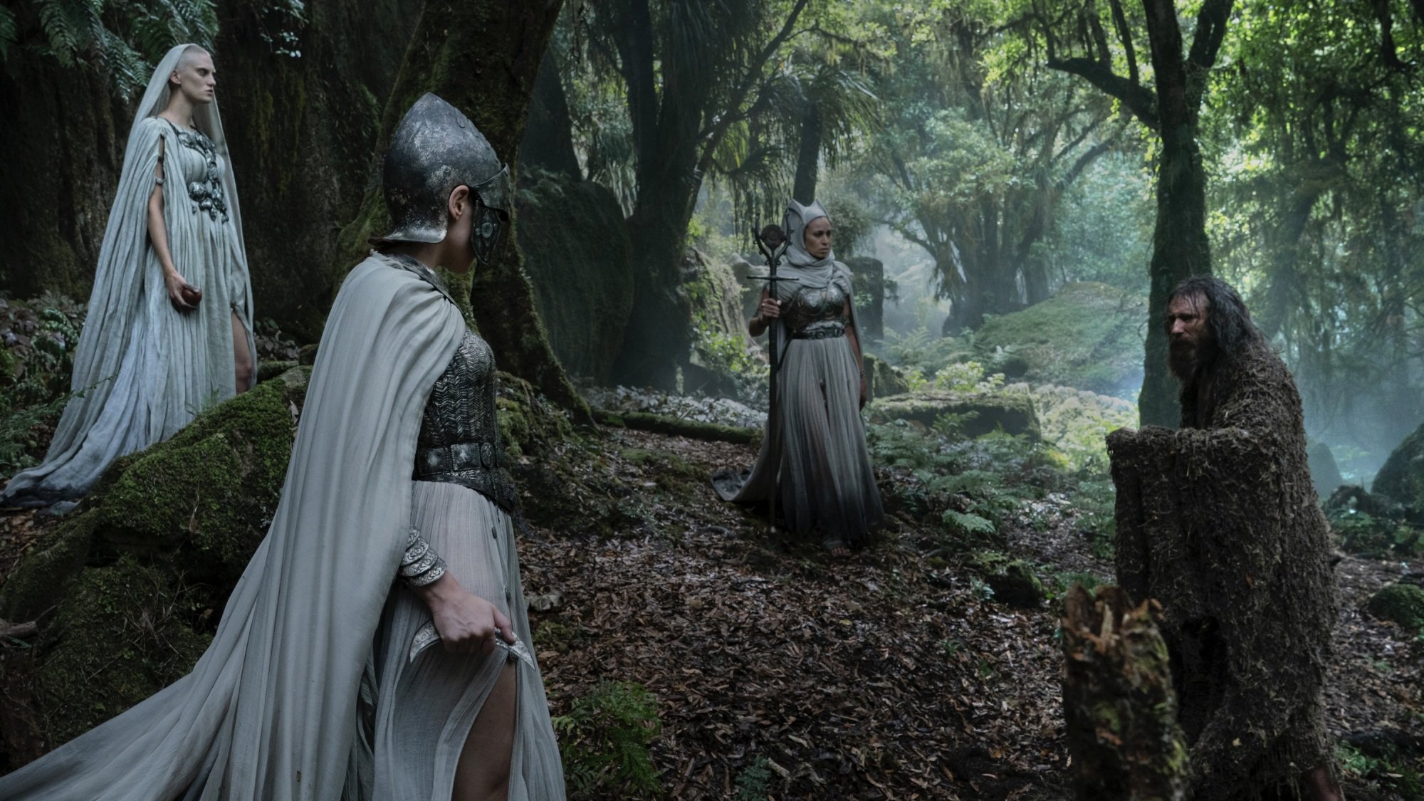 A tall man in a brown disheveled robe faces off against three imposing white-cloaked women in a forest.