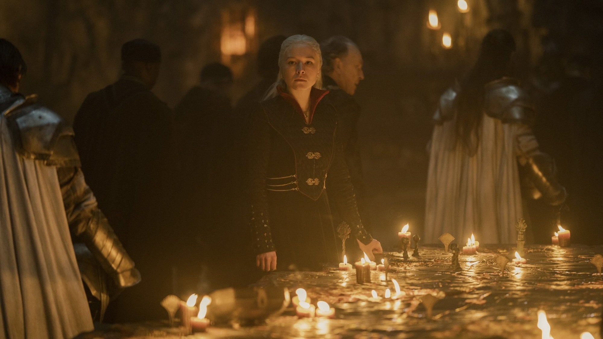 A woman with silver blonde hair in a black cloak stands at a war table covered in candles. Knights swarm behind her.