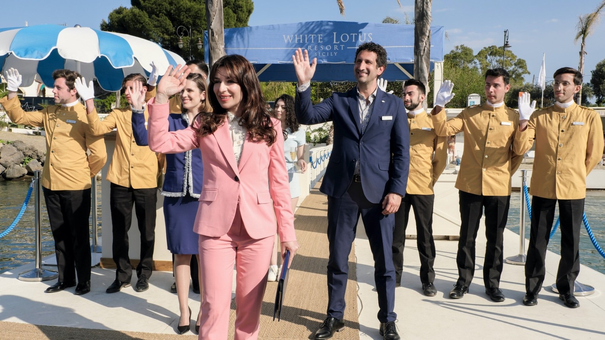 A woman in a pink suit and a group of hotel staff in yellow blazers stand on a dock, waving.