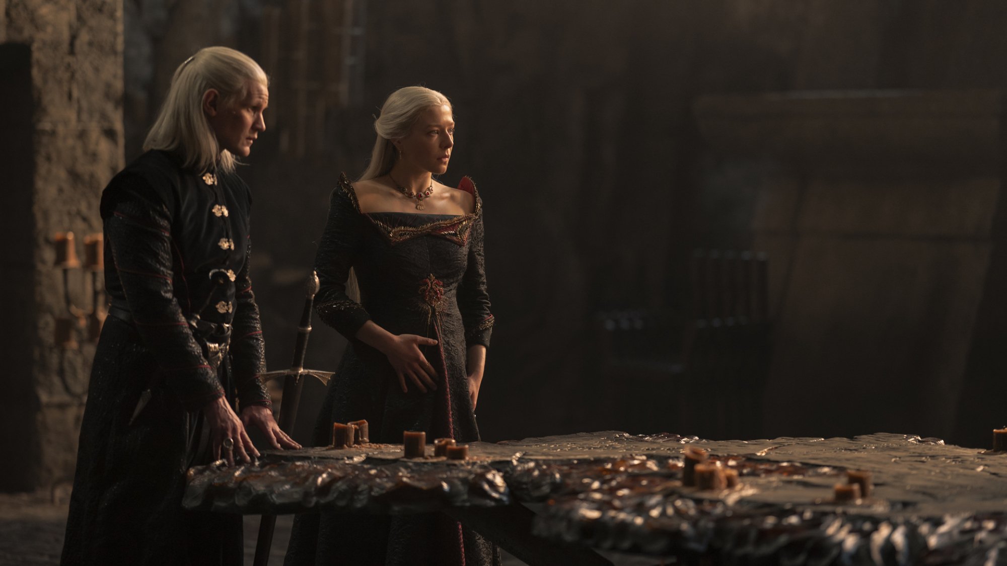 Two actors dressed in fantasy aristocratic costumes stand on front of a map table.