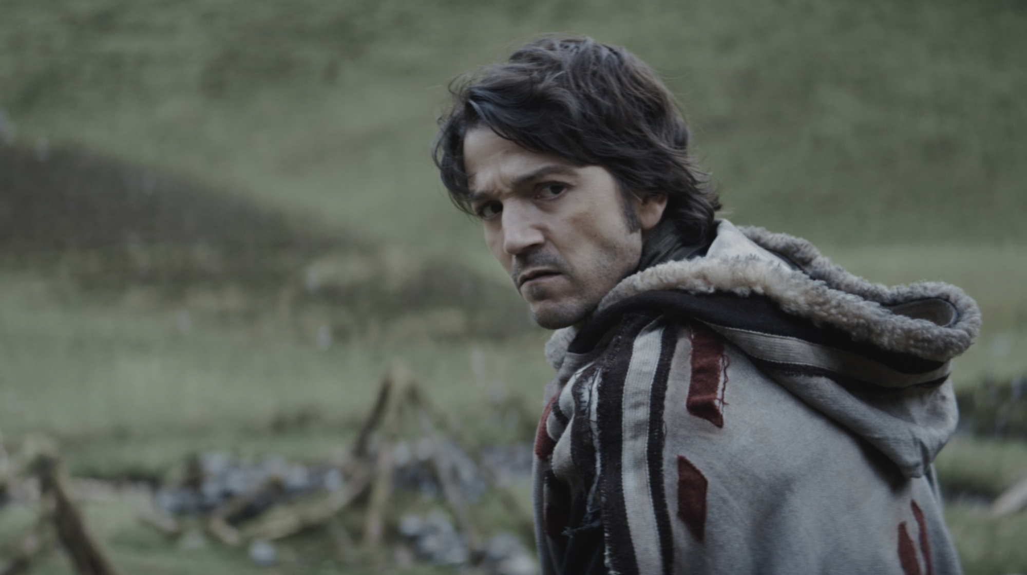 A dark-haired man walking outdoors in a hooded cloak looks serious.