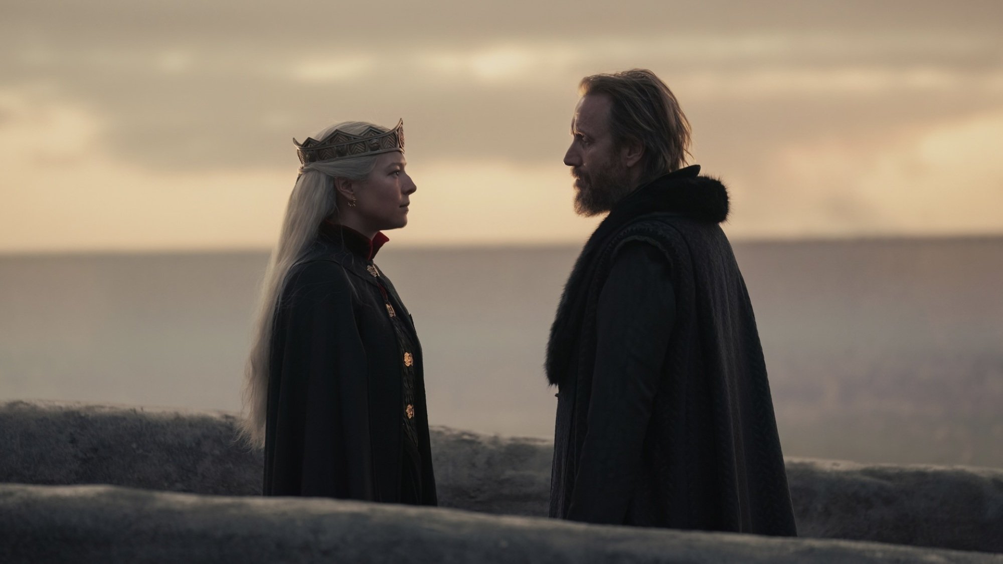 A woman with silver blonde hair in a crown and black cloak and a man in a black fur cloak stand on a stone path overlooking the ocean.