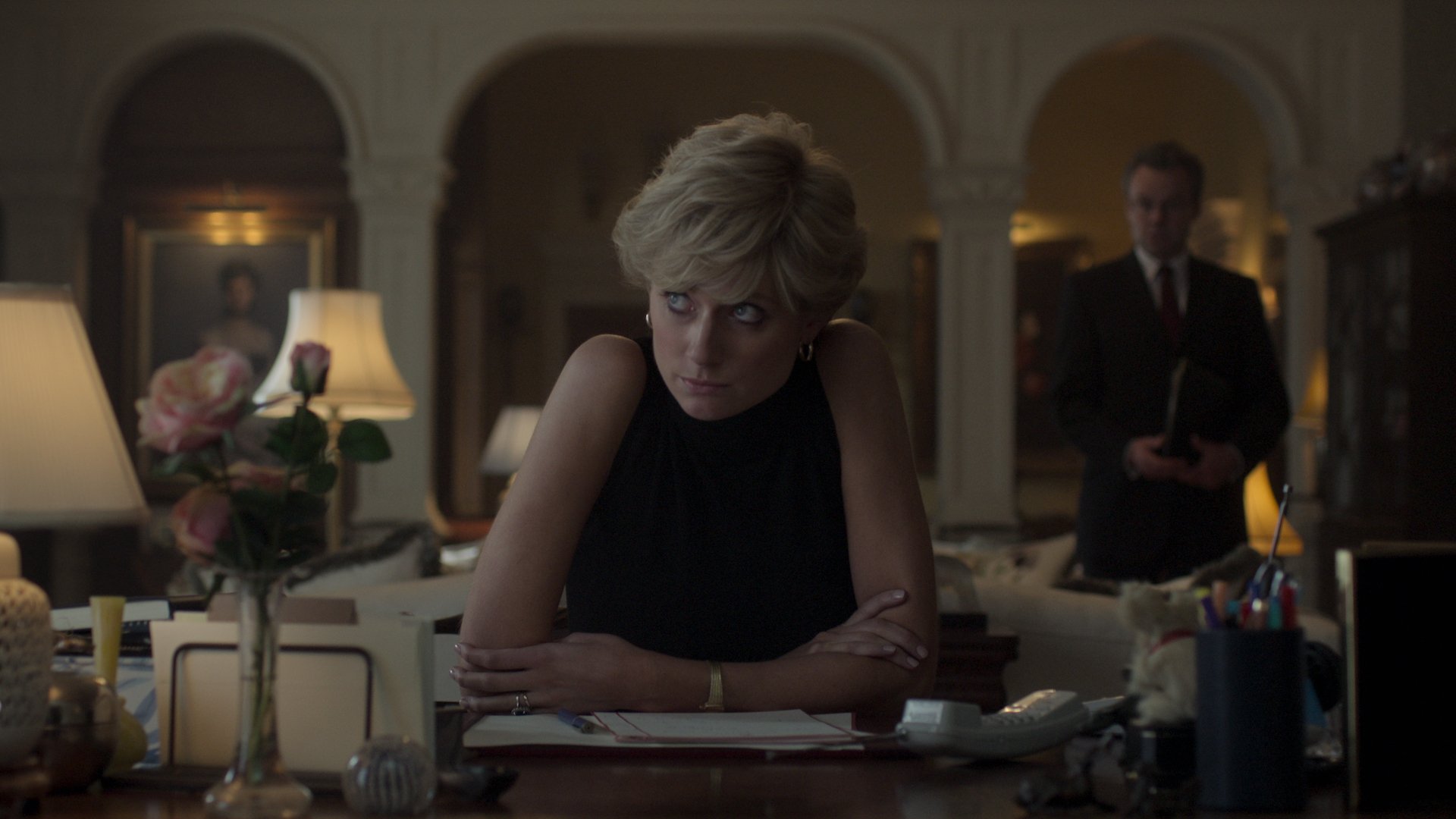 A woman in a black sleeveless top in an aristocratic lounge room sits at a desk looking stressed.