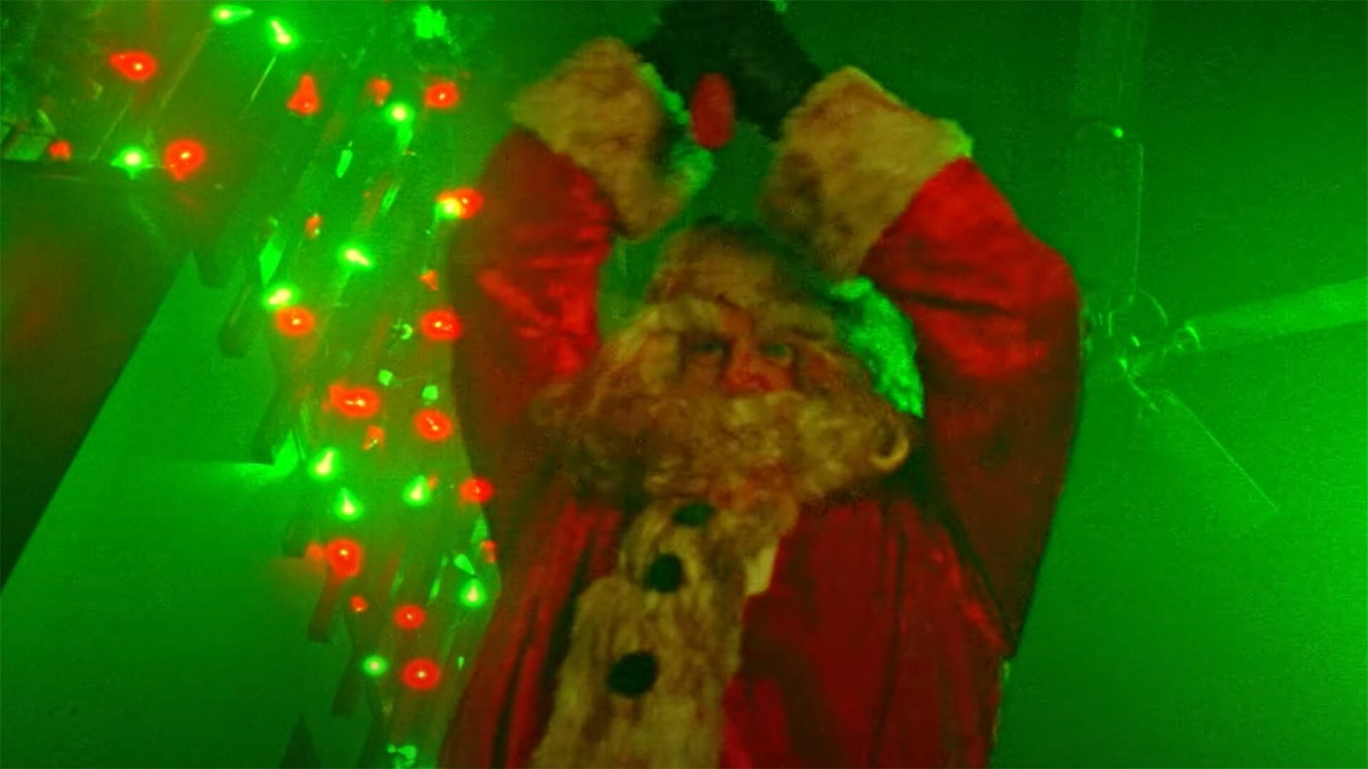 A man in a Santa Claus outfit with blood on his beard raises an axe over his head.
