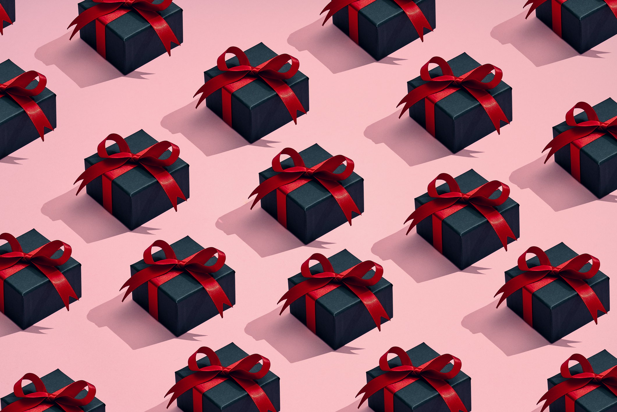 Repeating pattern of navy colored gift boxes with a red ribbon over a pink background.