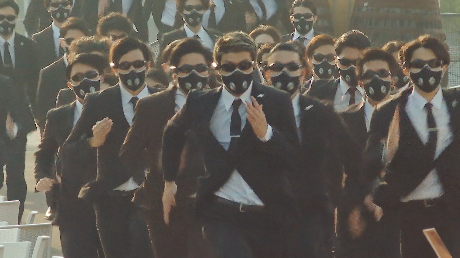 A group of men in black suits, with black face masks and sunglasses run towards the camera.