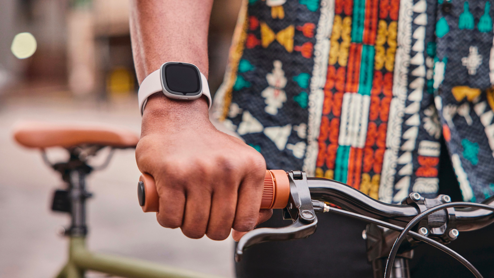 a close-up of a fitbit sense 2 on a person's wrist as they hold a bike's handlebar