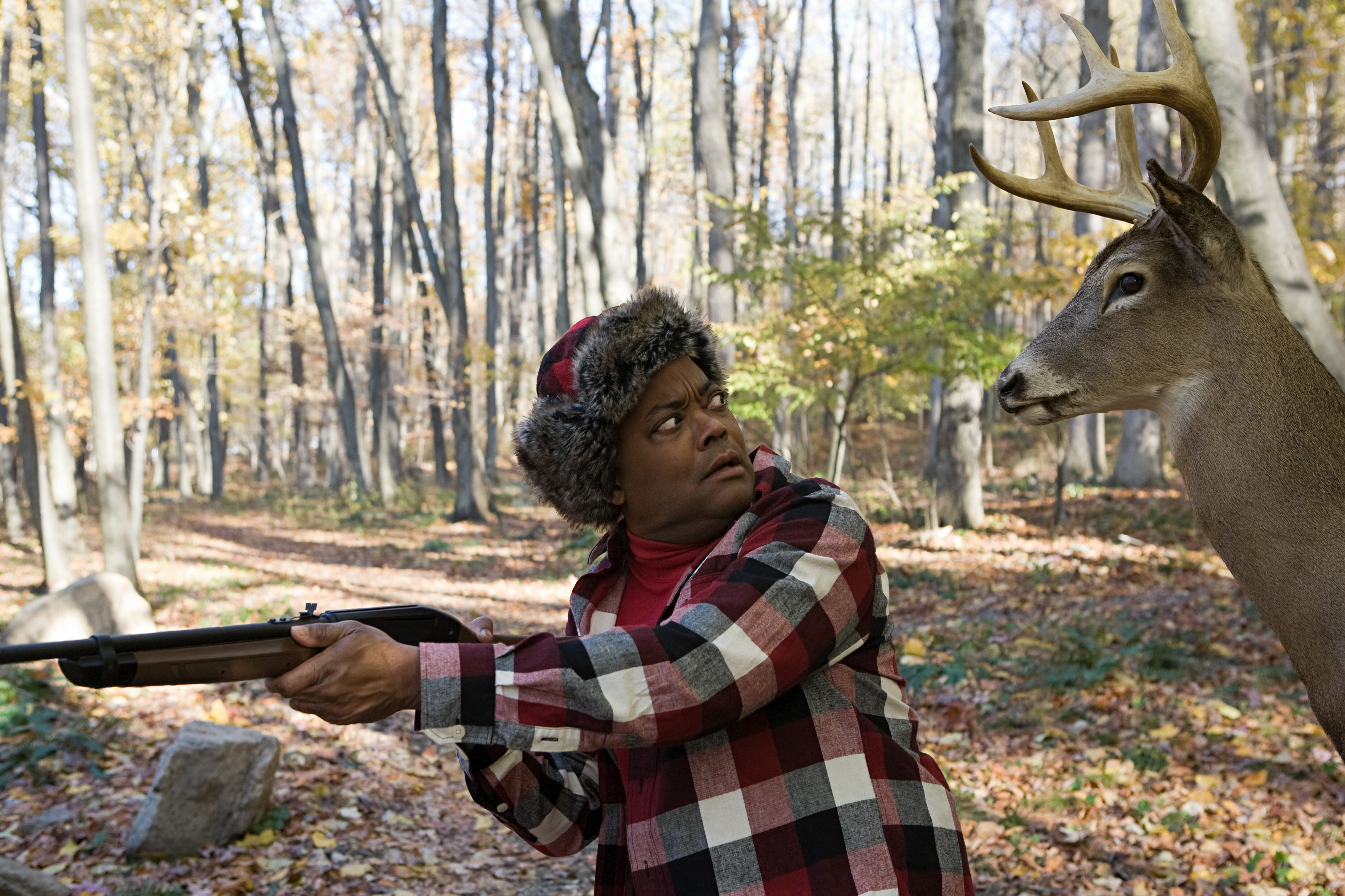 A man in plaid hunting clothes holds up a rifle. He is looking over his shoulder and making eye contact with a large deer.