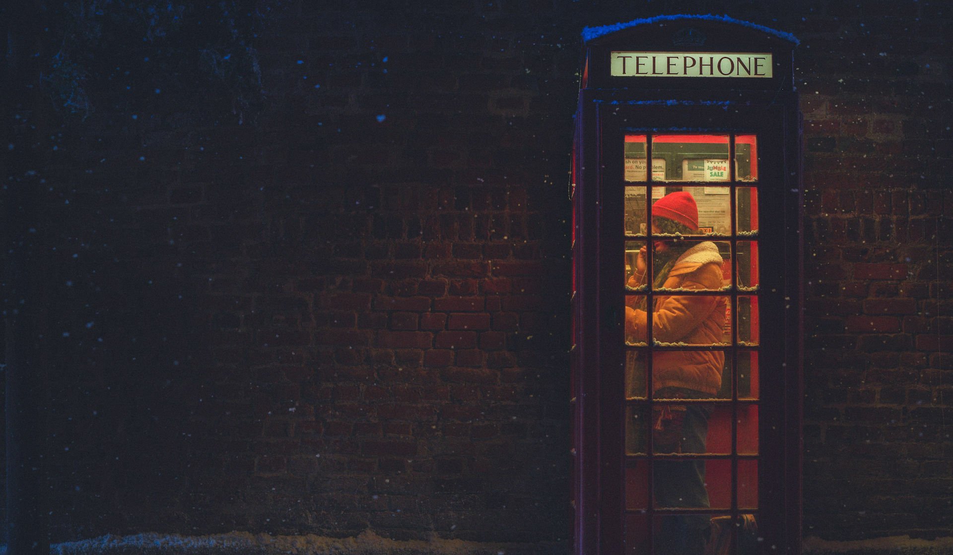 A girl with a red beanie stands in a British phone box at night in the snow.