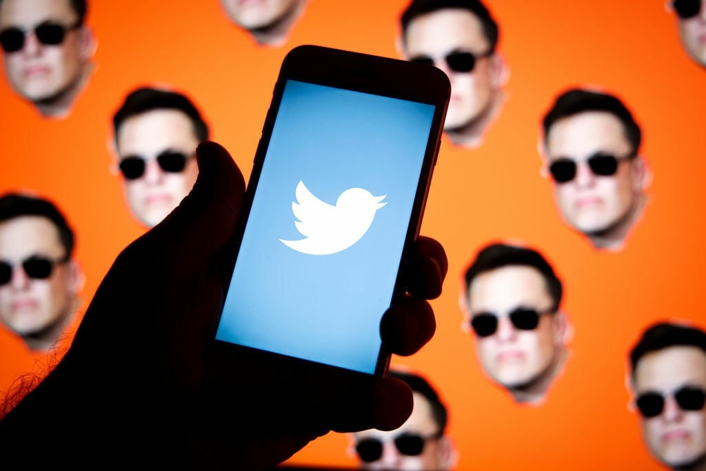 The Twitter logo on a smartphone, over a background of Elon Musk's heads.
