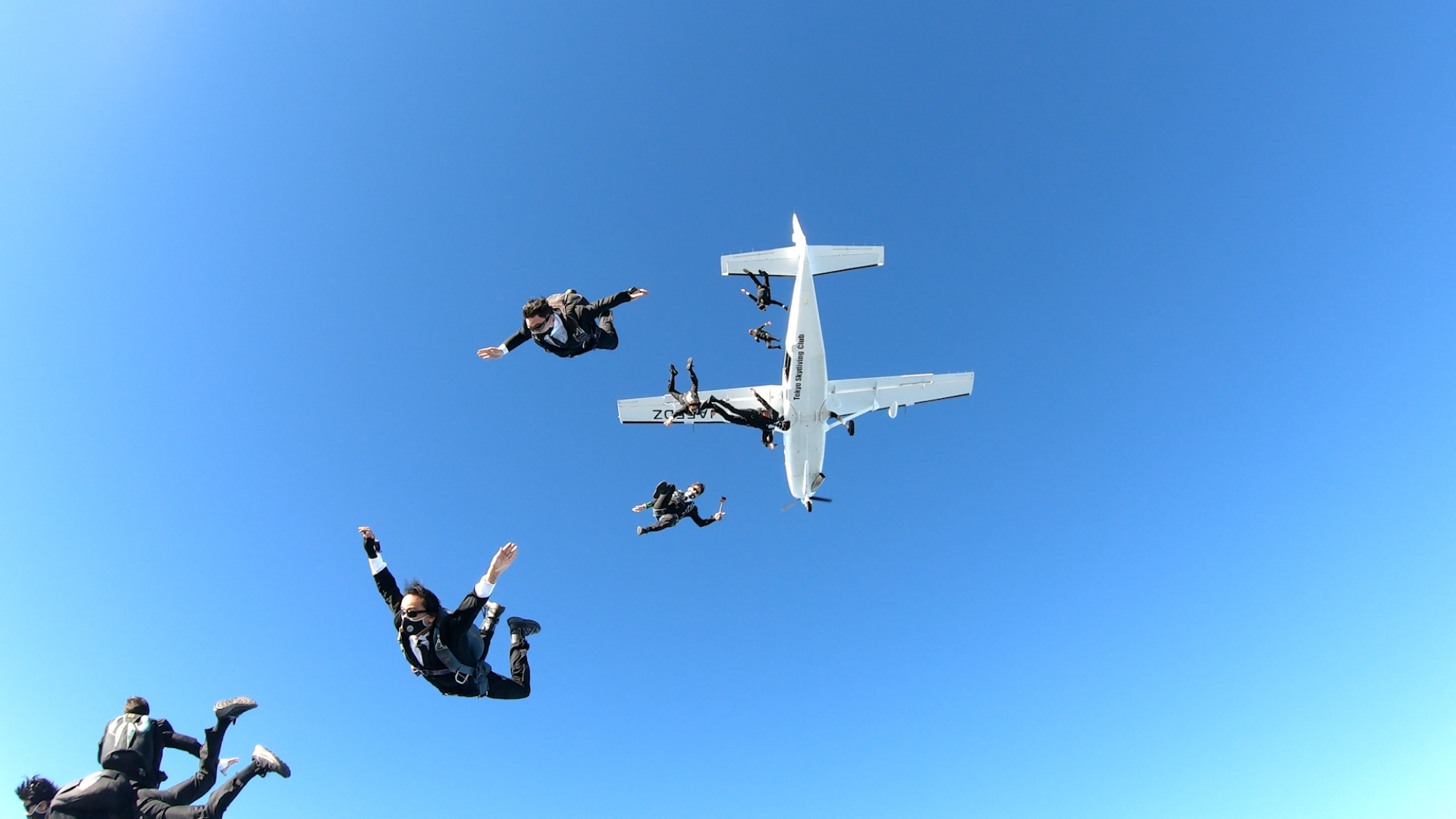 A group of men in dark suits and sunglasses dive from a plane in a blue sky, seen from below.