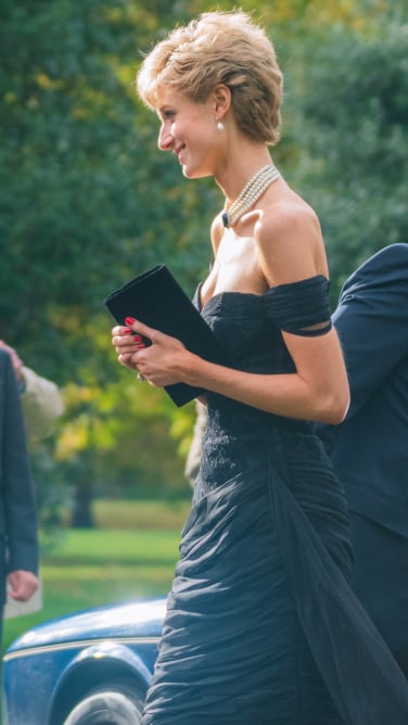 A woman in an off-the-shoulder black evening gown walks holding a clutch.