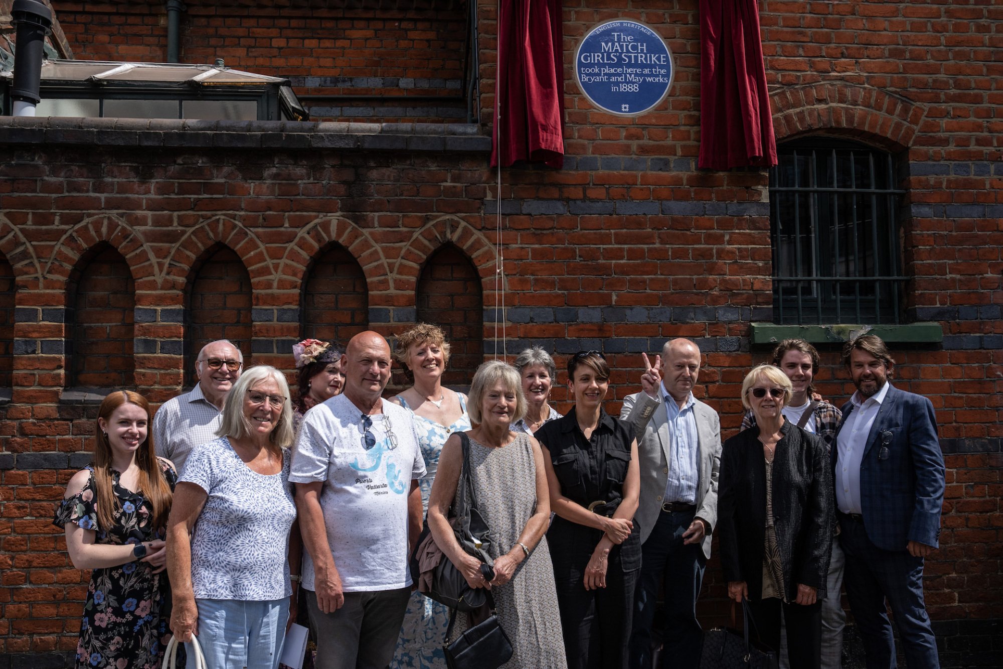 A group of people stand for a photograph together outside a brick wall with a blue plaque on it.