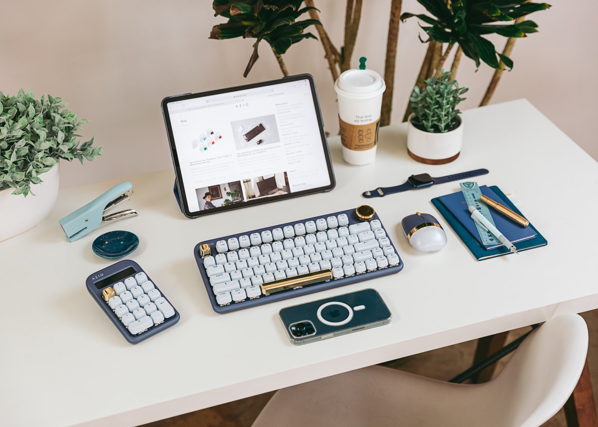 A work setup on a white desk consisting of an iPad and dark blue keyboard with gold accents, matching calculator. matching mouse, and a face down iPhone.
